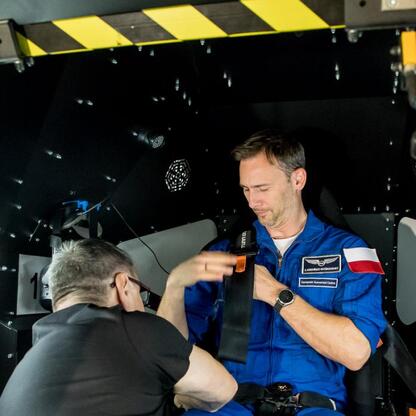 Astronauta Sławosz Uznański-Wiśniewski w symulatorze lotu kosmicznego, wspierany przez osobę w białym mundurze, w ramach misji IGNIS. (wygenerowano przez AI)