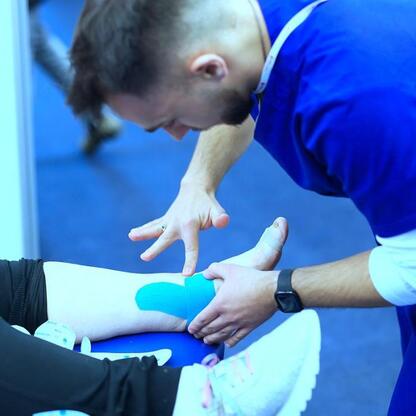 Zdjęcie przedstawia lekarza w niebieskim mundurze, który nakłada tapeczkę kinesiotape na kolano pacjenta w środowisku medycznym, prawdopodobnie Śląskim Stadionie. (wygenerowano przez AI)