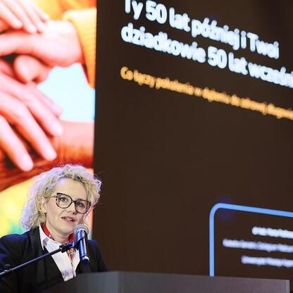 Na Śląsku, w III edycji Centralnego Klubu Pacjenta, kobieta przemawia na podium przed dużym ekranem z prezentacją o połączeniu pokoleń. (wygenerowano przez AI)