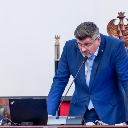 Na zdjęciu przedstawiono sesję Sejmiku Województwa Śląskiego, gdzie radny w niebieskim garniturze przemawia do mikrofonu. (wygenerowano przez AI)