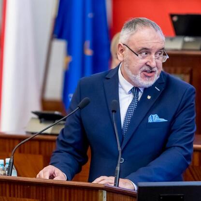 Mężczyzna w ciemnoniebieskim garniturze i krawacie stoi na podium, przemawiając w formalnym uroczystości w budynku administracyjnym. (wygenerowano przez AI)