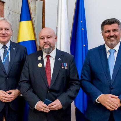 Trzech urzędników w mundurach z medalami stoi przed flagami, reprezentującymi UE i Śląsk, podczas sesji Sejmiku Województwa Śląskiego. (wygenerowano przez AI)