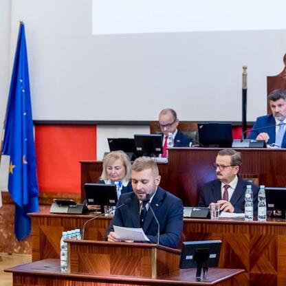 W trakcie sesji Sejmiku Województwa Śląskiego radny przemawia, za nim siedzący ludzie notują lub obserwują. (wygenerowano przez AI)