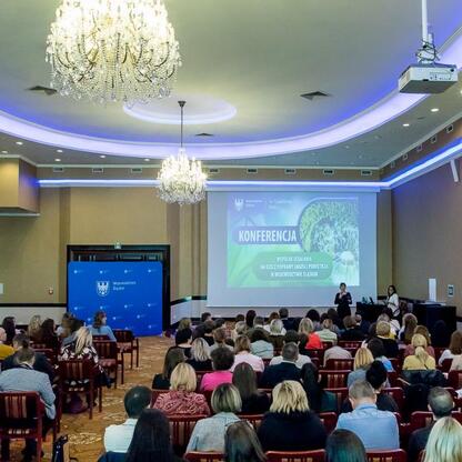 Konferencja o poprawie jakości powietrza w Śląsku, uczestnicy w czerwonych krzesłach, prezentacja na ekranie. (wygenerowano przez AI)