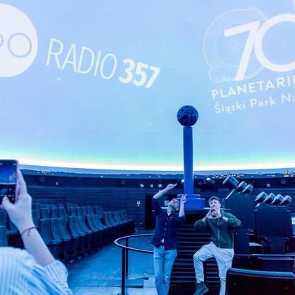 Wśród tłumu słuchaczy w Planetarium Śląskiego, widoczna jest osoba robiąca zdjęcie dwóch osób podczas koncertu Radio 357. (wygenerowano przez AI)