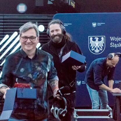 Zdjęcie przedstawia moment z transmisji Radiu 357 z Planetarium Śląskiego, gdzie osobę w słuchawkach i osobę z pudełkiem na tle logo Województwa Śląskiego. (wygenerowano przez AI)