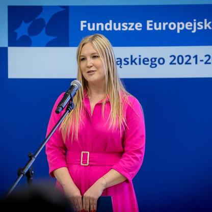 Joanna Bojczuk, członek Zarządu Województwa Śląskiego, przemawia na podium podczas uroczystości związanych z Europejskimi Funduszami 2021-2027. (wygenerowano przez AI)