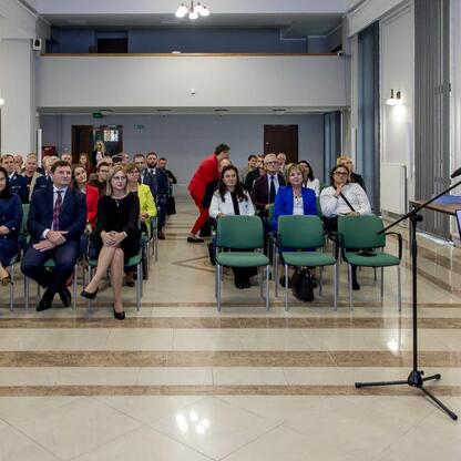 Widoczna jest formalna konferencja w Sali Konferencyjnej Urzędu Marszałkowskiego Województwa Śląskiego, gdzie kobieta w różowej sukience przemawia z podium, a słuchacze w czarnych garniturach siedzą w (wygenerowano przez AI)
