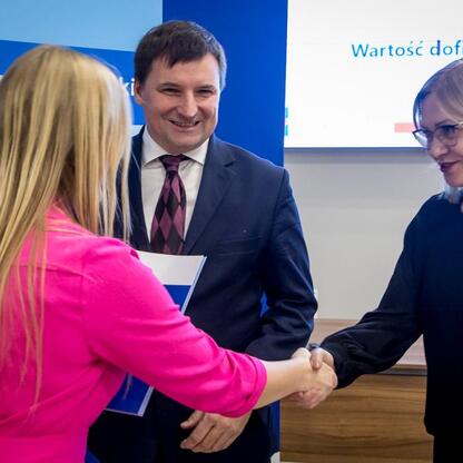 Dwie kobiety witały się na tle informacji o projekcie Funduszy Europejskich dla Śląska 2021-2027. (wygenerowano przez AI)