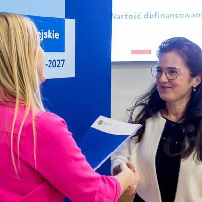 Dwie kobiety składają sobie gratulacje przed logo i tekstem funduszy europejskich dla Śląska 2021-2027. (wygenerowano przez AI)