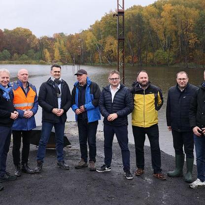 Grupa dziewięciu mężczyzn podczas spotkania wokół jeziora, omawiających reaktywację linii kolejowej Katowice-Jastrzębie. (wygenerowano przez AI)