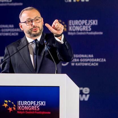 Mężczyzna w garniturze i okularach przemawia na podium podczas Europejskiego Kongresu Małych i Średnich Przedsiębiorstw w Katowicach. (wygenerowano przez AI)