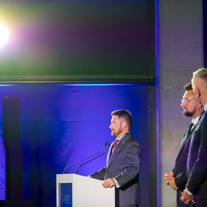 Mężczyzna w ciemnym garniturze przemawia na podium podczas Europejskiego Kongresu Małych i Średnich Przedsiębiorstw w Katowicach. (wygenerowano przez AI)