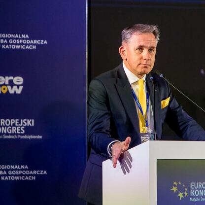 Mężczyzna w ciemnym garniturze i żółtym krawacie przemawia na podium podczas Europejskiego Kongresu Małych i Średnich Przedsiębiorstw w Katowicach. (wygenerowano przez AI)
