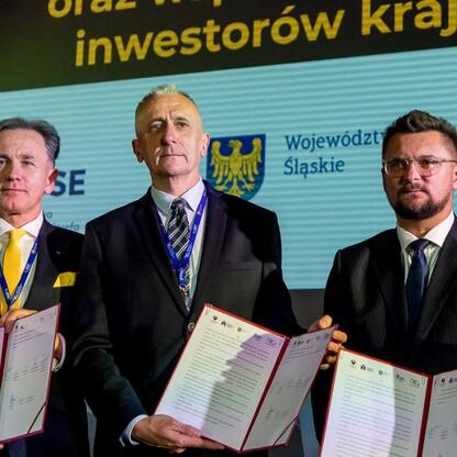 Trzech mężczyzn w strojach formalnych przed logo Województwa Śląskiego i KSSE, trzymają dokumenty, symbolizujące współpracę między instytucjami. (wygenerowano przez AI)