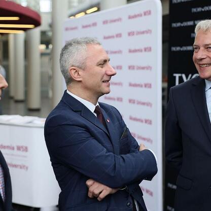 Trzech mężczyzn w strojach formalnych przed logo WSB University, dyskutujący o bezpieczeństwie i odporności cywilnej i gospodarczej. (wygenerowano przez AI)