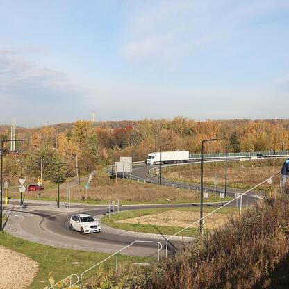 Nowo wybudowana droga w ramach Trasy N-S, łącząca A4 z DTŚ, z widocznym ruchem samochodów i pieszymi na tle zielonej i urbanistycznej zabudowy. (wygenerowano przez AI)