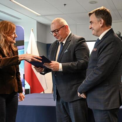 Trzy osoby w strojach formalnych uczestniczą w ceremonii wymiany dokumentów, związanej z dofinansowaniem zakupu nowych pociągów dla Kolei Śląskich. (wygenerowano przez AI)
