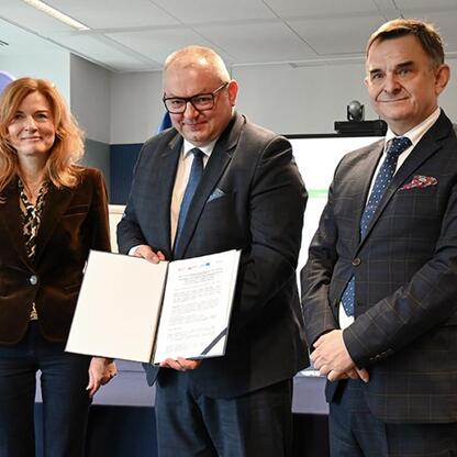 Trzy osoby w formalnym stroju biznesowym stoją przed logo CUPT, uczestnicząc w ceremonii podpisania umów finansowania zakupu nowych pociągów dla Kolei Śląskich. (wygenerowano przez AI)