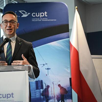 Mężczyzna przemawia na podium w Centrum Unijnych Projektów Transportowych, podkreślając znaczenie finansowania nowych pociągów dla Kolei Śląskich. (wygenerowano przez AI)