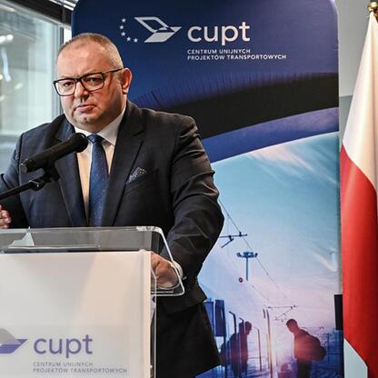 Mężczyzna w garniturze przemawia na podium z mikrofonem, przedstawiając projekt finansowania nowych pociągów dla Kolei Śląskich. (wygenerowano przez AI)