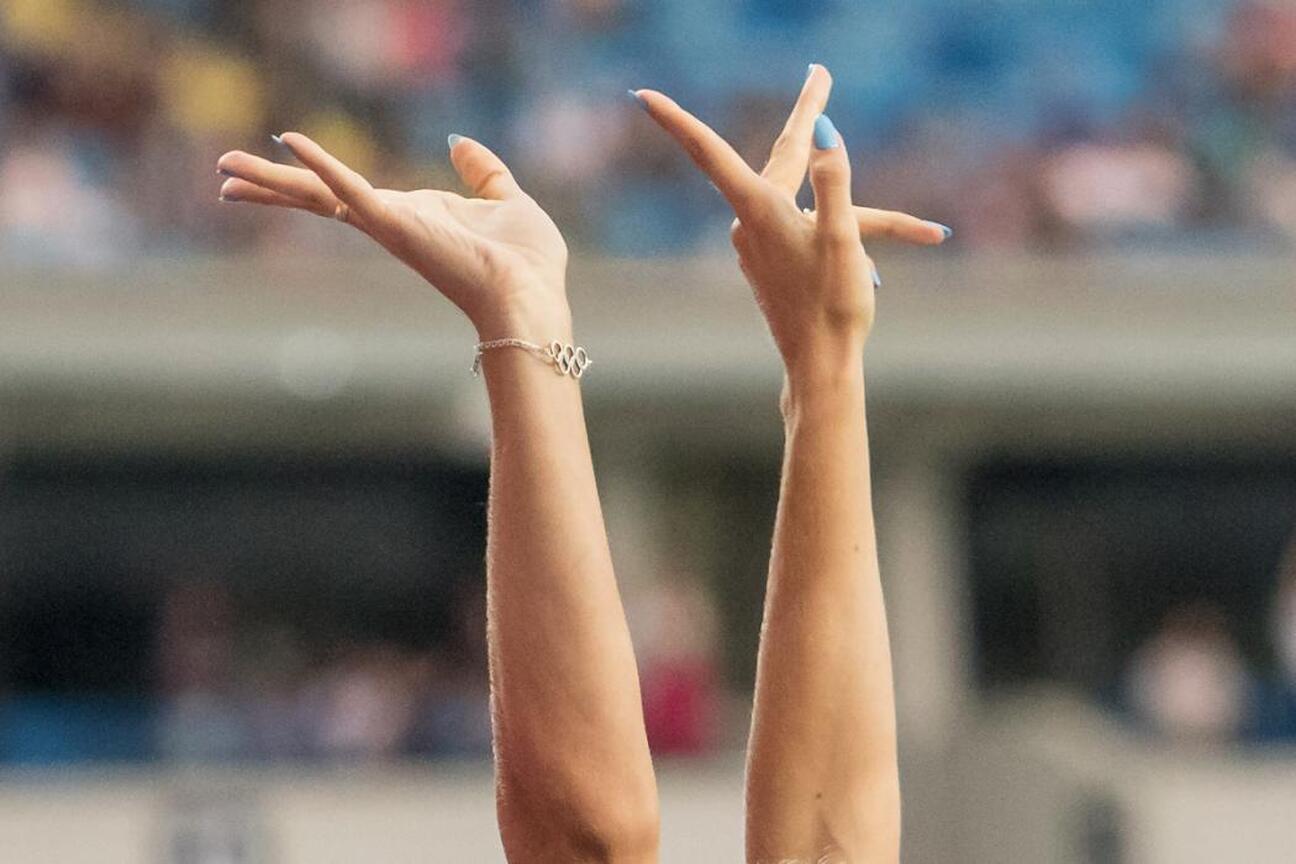 Zdjęcie przedstawia ręce z bransoletą z olimpijskimi pierścieniami, udekorowane niebieskimi paznokciami, podniesione w "V" kształt, na tle rozmytego stadionu. (wygenerowano przez AI)
