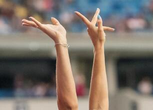 Zdjęcie przedstawia ręce z bransoletą z olimpijskimi pierścieniami, udekorowane niebieskimi paznokciami, podniesione w "V" kształt, na tle rozmytego stadionu. (wygenerowano przez AI)