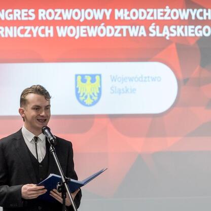 III Kongres Rozwojowy Młodzieżowych Drużyn Pożarniczych Województwa Śląskiego, Superauto.pl Stadium. Młody człowiek w garniturze przemawia na podium, trzymając mikrofon i folder. (wygenerowano przez AI)