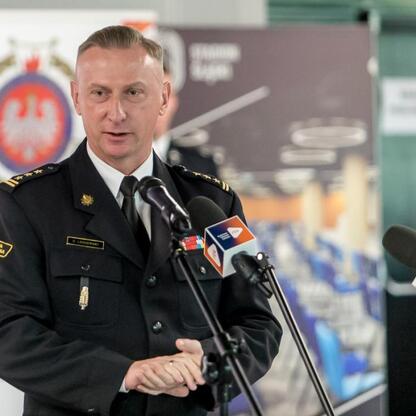 Na podium w czarnym mundurze z złotymi wstęgami stoi przed mikrofonami, prawdopodobnie przemawiający na Kongresie Rozwojowym Młodzieżowych Drużyn Pożarniczych Województwa Śląskiego. (wygenerowano przez AI)