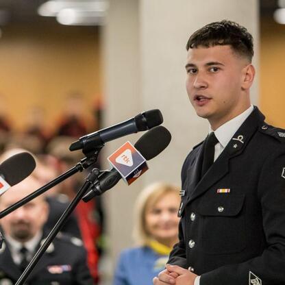 Podczas trzeciej edycji Kongresu Rozwojowego Młodzieżowych Drużyn Pożarniczych Województwa Śląskiego na Superauto.pl Stadionu Śląskiego młody strażak przemawia do zgromadzonej publiczności. (wygenerowano przez AI)