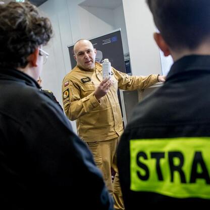 Na zdjęciu przedstawiono scenę z Kongresu Rozwojowego Młodzieżowych Drużyn Pożarniczych Województwa Śląskiego, gdzie uczestnik w mundurze demonstruje używanie strażackiego sprzętu. (wygenerowano przez AI)