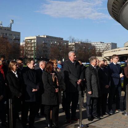 Grupa oficjalnych osób w strojach formalnych stoi przed Katowickim Spodekiem, odbierając nowe hybrydowe radiowozy. (wygenerowano przez AI)