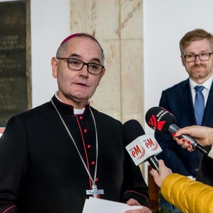 Biskup w czerni i czerwieni przemawia na konferencji prasowej, otoczony dziennikarzami z logo 