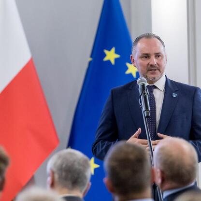 Mężczyzna w ciemnym garniturze i krawacie przemawia do mikrofonu, tło: flagi UE i Polski, Silesia. (wygenerowano przez AI)
