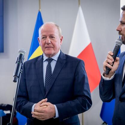Dwóch mężczyzn w strojach formalnych stoi przed flagami, prawdopodobnie podczas ceremonii oficjalnej. (wygenerowano przez AI)