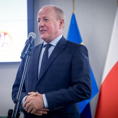 Mężczyzna w ciemnym garniturze i krawacie stojący na podium z mikrofonem, przemawiający na ceremonii wręczenia nagród wojewódzkich. (wygenerowano przez AI)