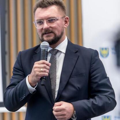 Mężczyzna w ciemnym garniturze i okularach przemawia do mikrofonu na formuлярnej imprezie, związany z zarządzaniem lub rozwijaniem Portu Lotniczego Katowice. (wygenerowano przez AI)