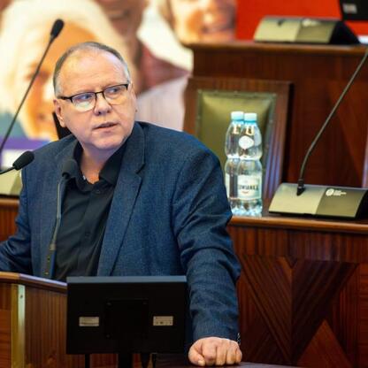 Mężczyzna w ciemnym garniturze siedzi przy stole, mikrofony i podium wskazują na rozmowę lub wystąpienie. (wygenerowano przez AI)