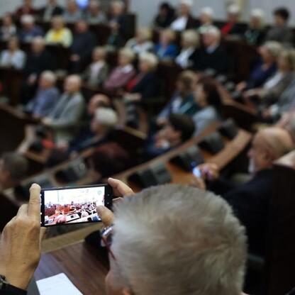 Zwiedzanie pierwszej edycji Śląskiego Konwentu Rad Seniorów w Sali Sejmu Śląskiego. Osoba w pierwszym rzędzie zapisuje na telefonie scenę z udziałem wielu słuchaczy. (wygenerowano przez AI)