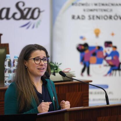 Podczas pierwszej edycji Śląskiego Konwentu Rad Seniorów w Sali Sejmu Śląskiego kobieta przemawia na podium, podkreślając znaczenie głosu seniorów w polityce regionalnej. (wygenerowano przez AI)