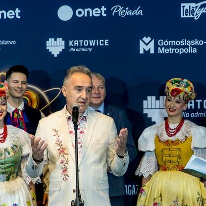 Mężczyzna w białym garniturze z kwiatowym haftem przemawia na podium, otoczony ludźmi w tradycyjnych strojach polskich. (wygenerowano przez AI)