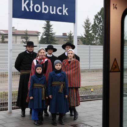 Zdjęcie przedstawia grupę ludzi w tradycyjnych strojach polskich, stojących na peronie stacji kolejowej Kłobuck, podczas testowego przejazdu nowego pociągu Impuls 2. (wygenerowano przez AI)
