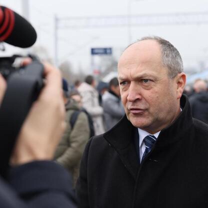 Na zdjęciu: Mężczyzna w czarnym garniturze rozmawia z operatorem kamery w stacji kolejowej. (wygenerowano przez AI)