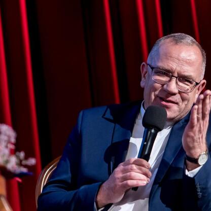 Mężczyzna w ciemnoniebieskim garniturze i okularach, mówiący do mikrofonu, podczas ceremonii wręczenia nagród Marszałkowskich w Sosnowcu. (wygenerowano przez AI)