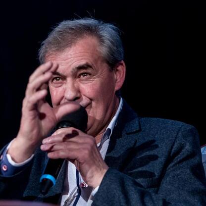 Mężczyzna w czarnej kamizelce i białej koszuli siedzi przy stole, gestykuluje, prawdopodobnie w trakcie panelu dyskusyjnego lub ceremonii w Kinoteatrze 