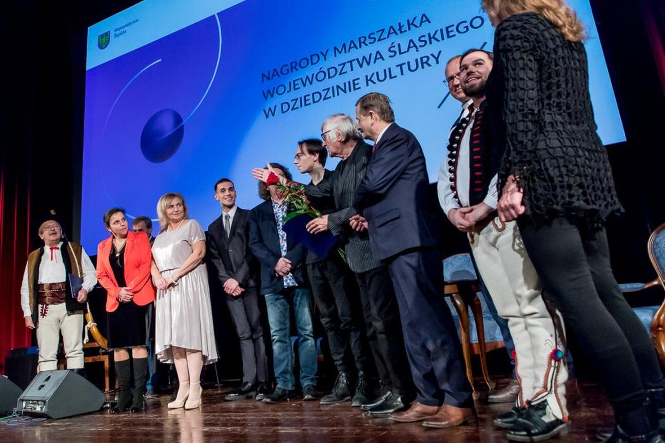Mężczyzna w tradycyjnym stroju trzyma instrument, uśmiechnięta kobieta w czarnej sukni obok. Uroczystość wręczenia nagród Marszałka Województwa Śląskiego w Kinoteatrze Rialto w Katowicach. (wygenerowano przez AI)