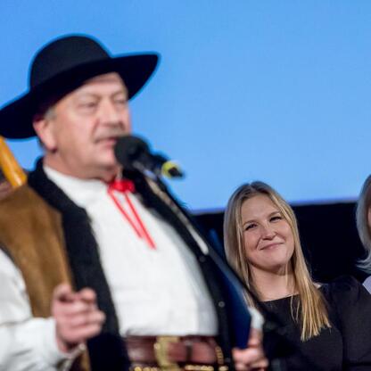 Mężczyzna w tradycyjnym stroju trzyma instrument, uśmiechnięta kobieta w czarnej sukni obok. Uroczystość wręczenia nagród Marszałka Województwa Śląskiego w Kinoteatrze Rialto w Katowicach. (wygenerowano przez AI)