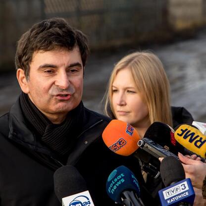 Mężczyzna w czarnym płaszczu przemawia do dziennikarzy, omawiając usuwanie niebezpiecznych odpadów w Katowicach-Szopienicach. (wygenerowano przez AI)