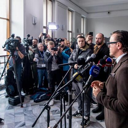 Widoczna jest konferencja prasowa w marبلowym korytarzu z dużymi oknami, gdzie dziennikarze ze sprzętem nagrywającym rozmawiają z mężczyzną w brązowym garniturze, reprezentującym rząd lub przemysł. (wygenerowano przez AI)