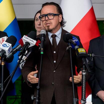 Fotografia przedstawia formalną konferencję prasową, gdzie mężczyzna w brązowym garniturze i okularach przesługuje mowę wokół mikrofonów różnych mediów, w tle flagi i wskazuje na rozmowę o przyszłości (wygenerowano przez AI)